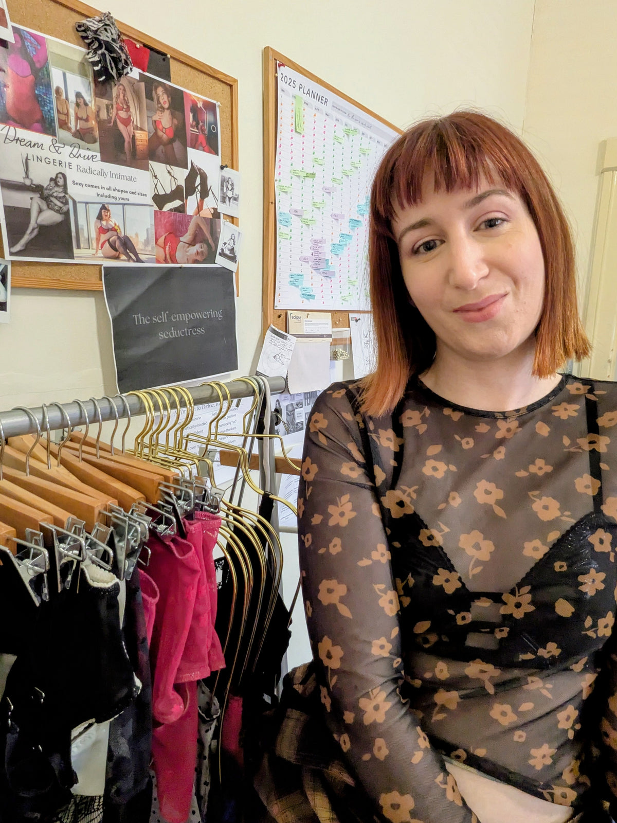 A woman with reddish-brown hair stands next to a clothing rack in a boutique, wearing a sheer black long-sleeve top with a tan floral pattern over a black bralette. The background features a clothing display rack with various garments on wooden hangers and a corkboard with photos and notes.