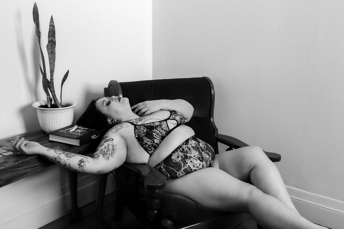 A black and white lifestyle photograph of a tattooed woman lying on a dark wooden armchair, wearing a patterned two-piece swimsuit. A potted snake plant sits on a side table next to her.